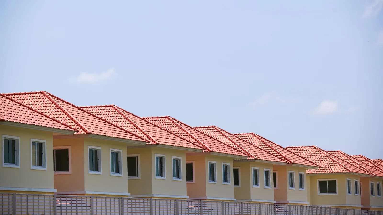 A line of identical suburban houses with red roofs and beige exteriors, perfectly aligned under a clear blue sky.