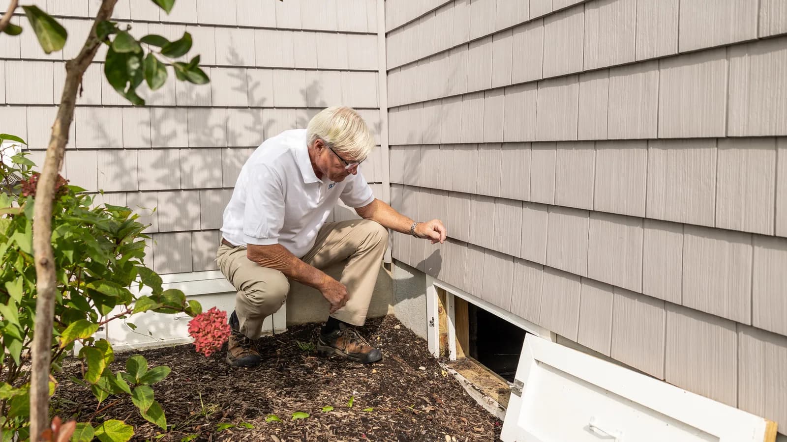 A person crouching beside an open exterior crawl-space access hatch