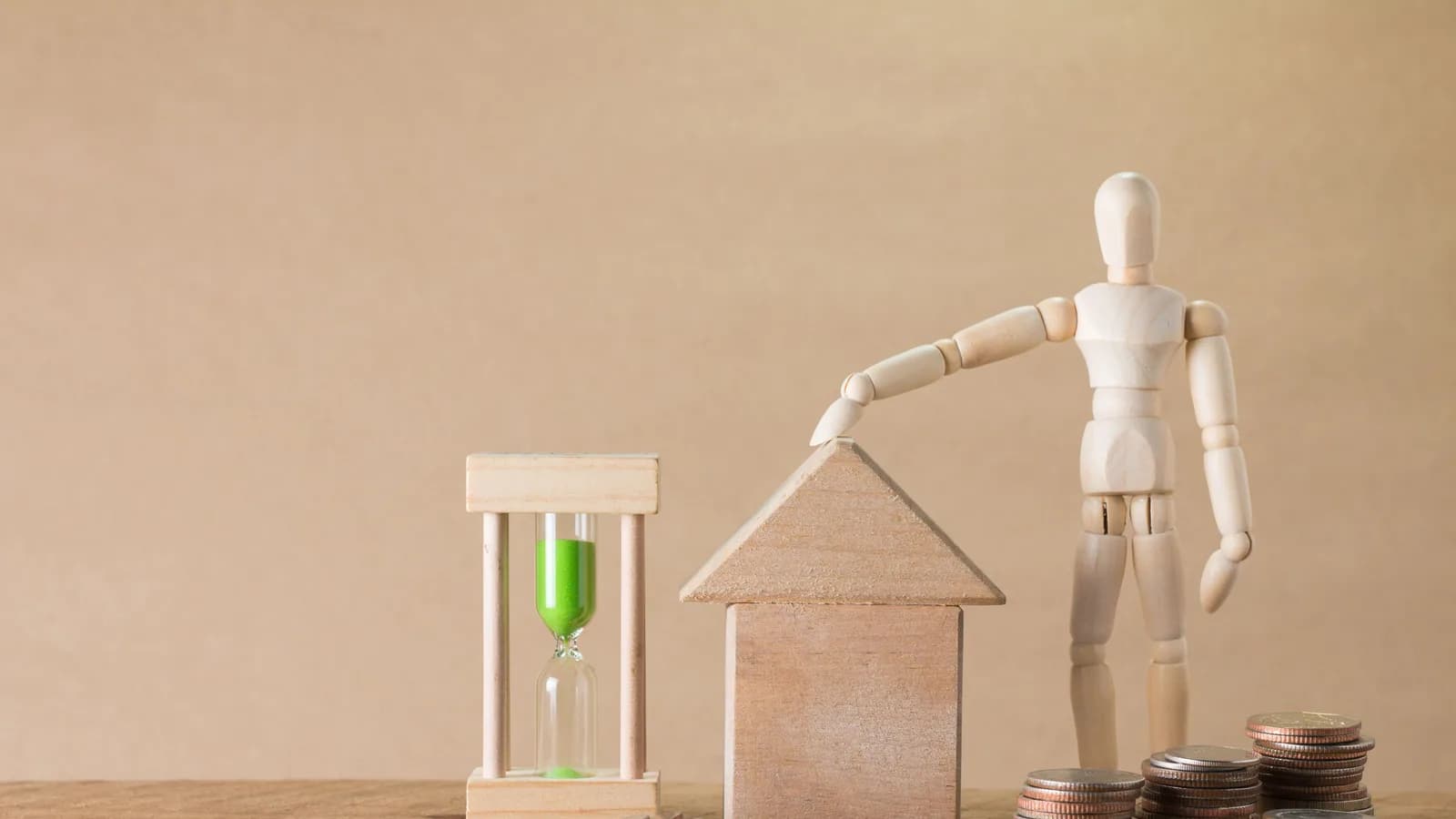 Wooden mannequin with house model, coins, and hourglass on a wooden table, symbolizing time and financial growth.