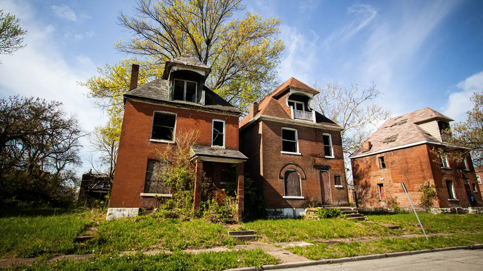 Three abandoned brick houses in a suburban neighborhood under a clear blue sky.