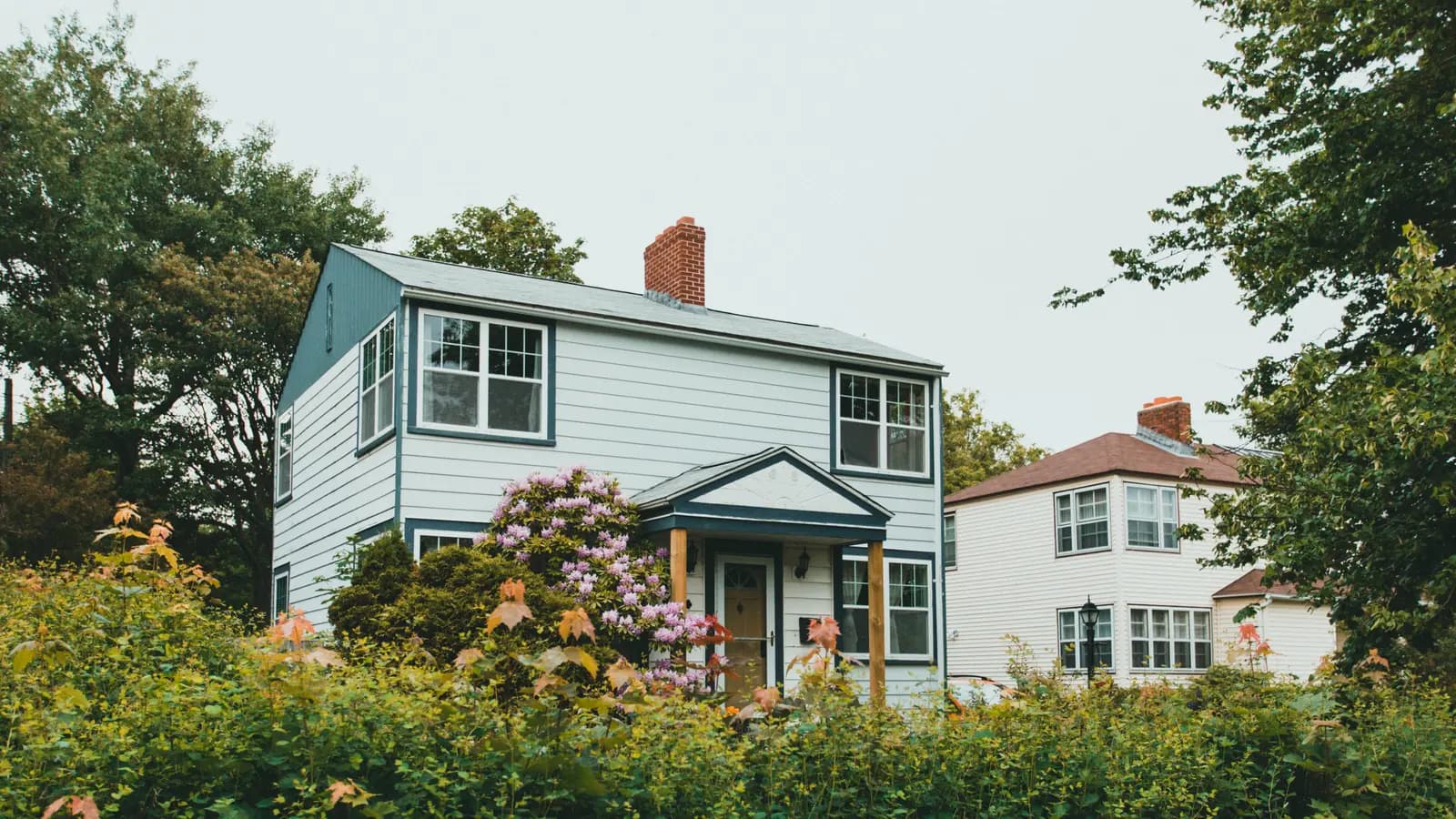 Charming two-story wooden house surrounded by lush greenery and vibrant flowers in a suburban neighborhood.