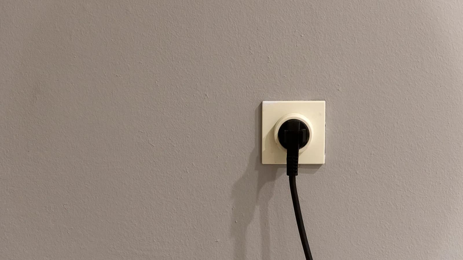 A black plug inserted into a white wall socket on a plain grey wall.
