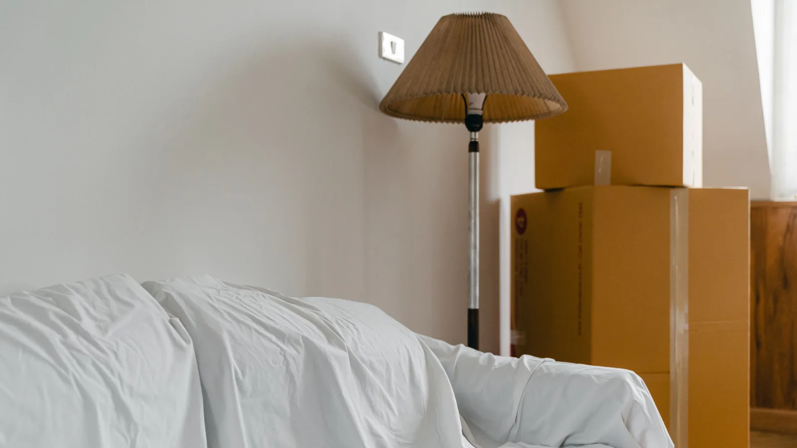 Covered sofa, floor lamp, and packed cardboard boxes in the light-filled room of a new home.