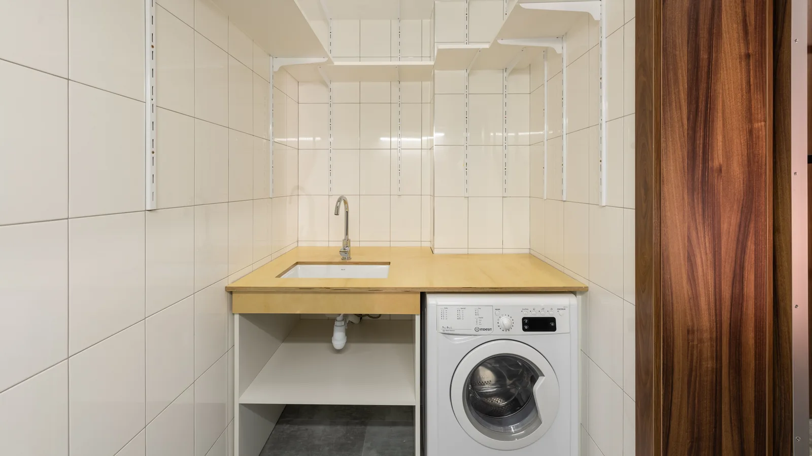 Compact laundry room with a washing machine, wooden counter and sink, and open wall shelves; no dryer or baskets