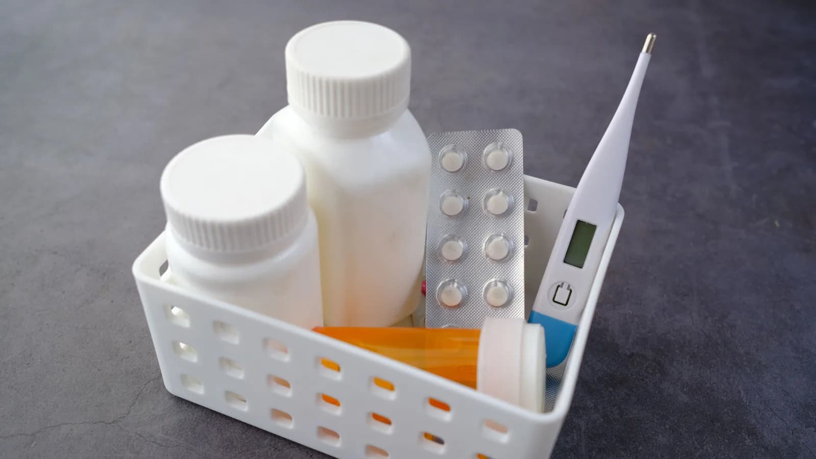 Two medicine bottles, a blister pack, an orange prescription bottle, and a digital thermometer in a white basket