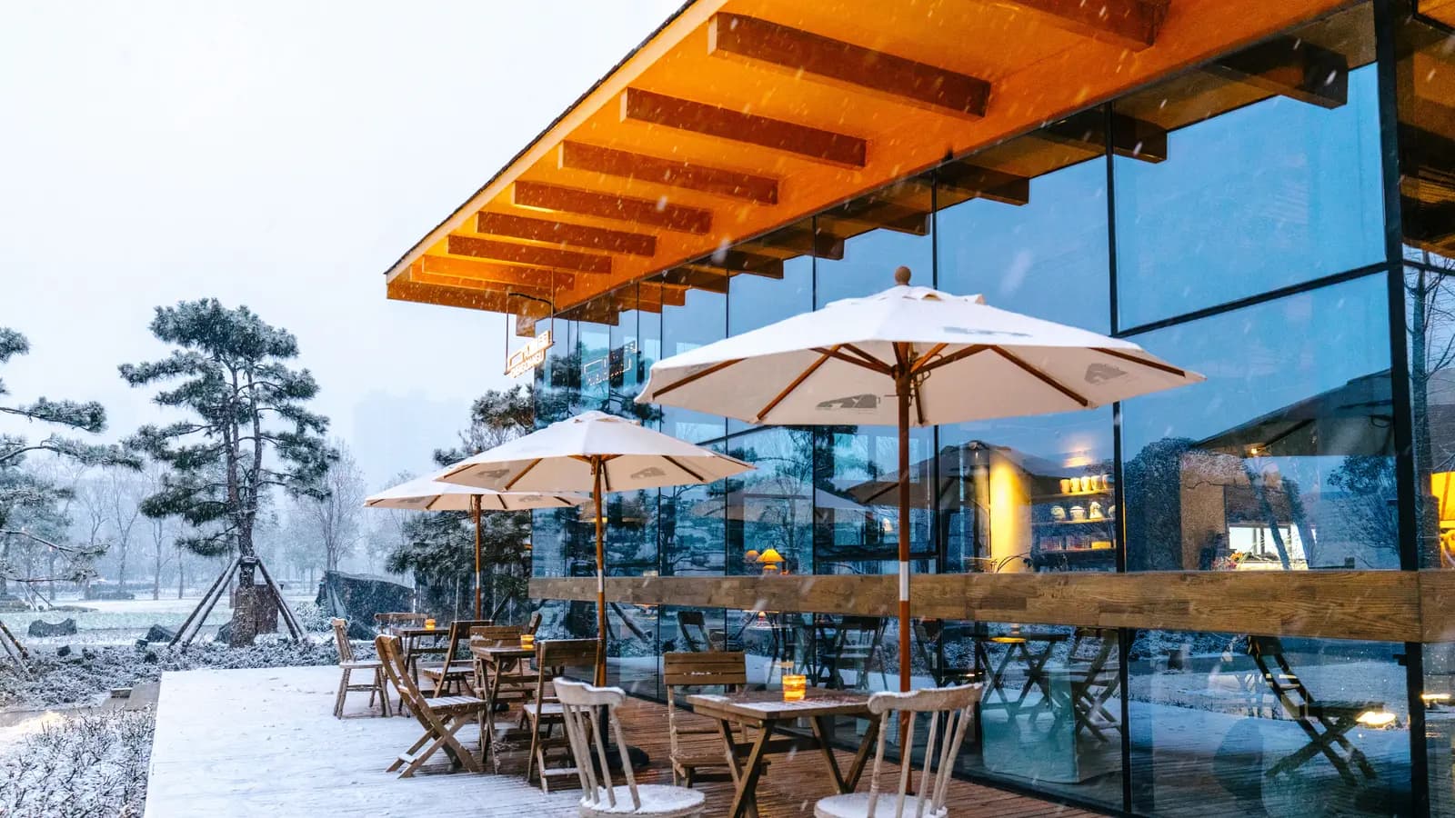 Wood patio tables, chairs, and umbrellas outside a glass building during snowfall
