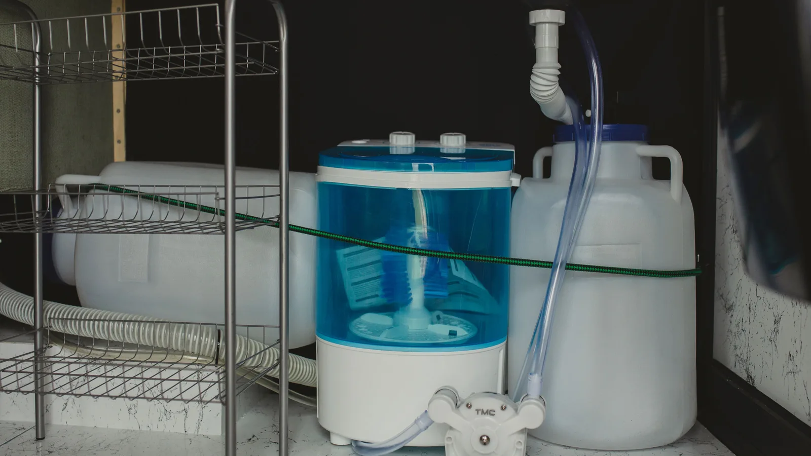 Compact travel-size washer in a storage area with plastic containers and tubing system.