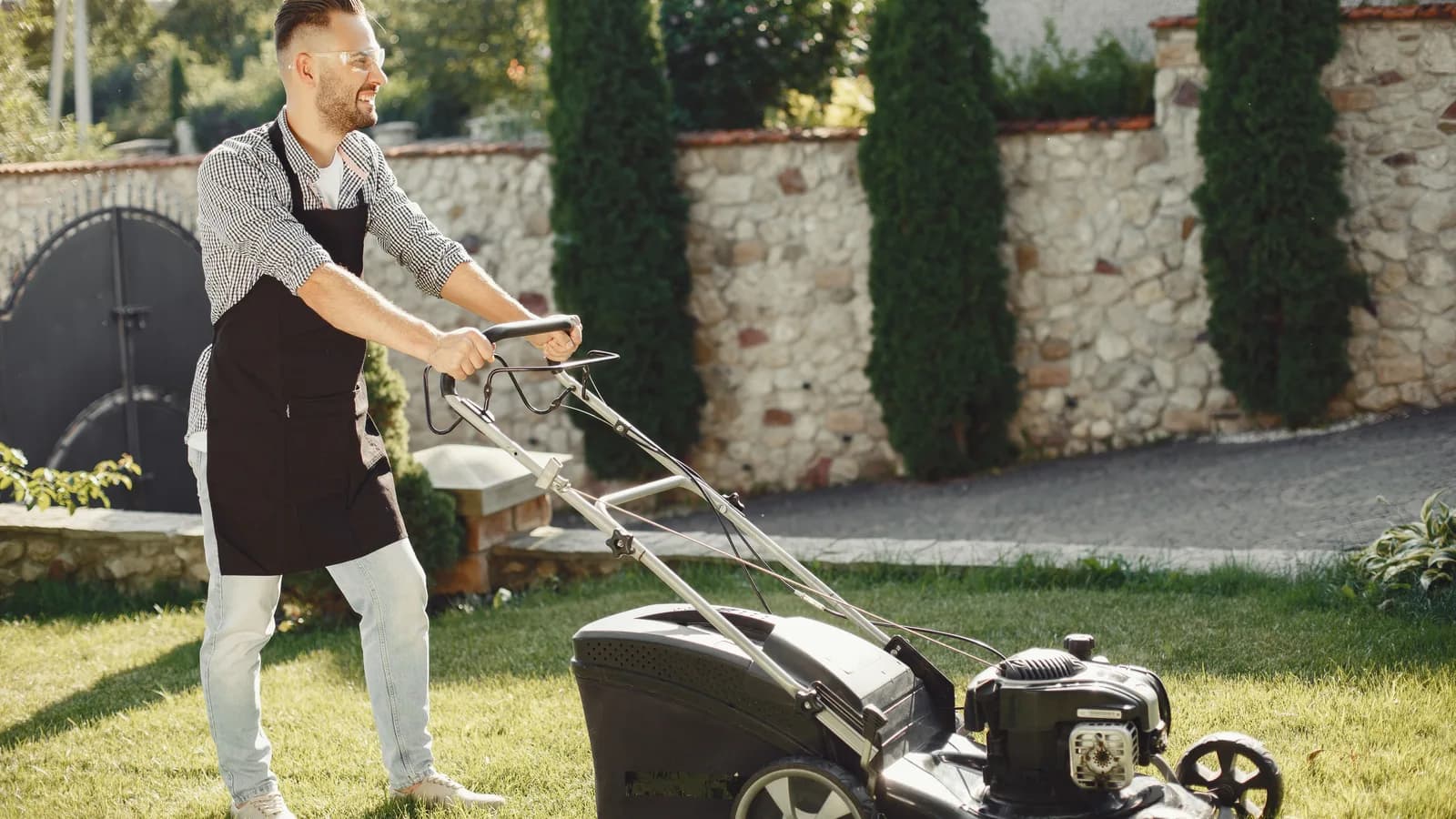 Man pushing a walk-behind lawn mower across a sunny residential lawn