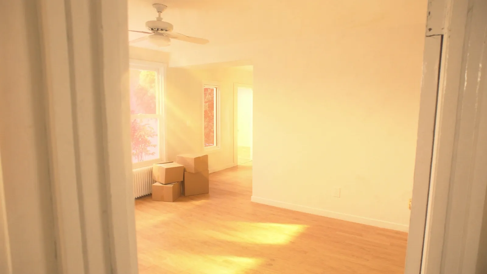 Empty sunlit living room of a starter home with a few stacked cardboard moving boxes and warm morning light on hardwood floors
