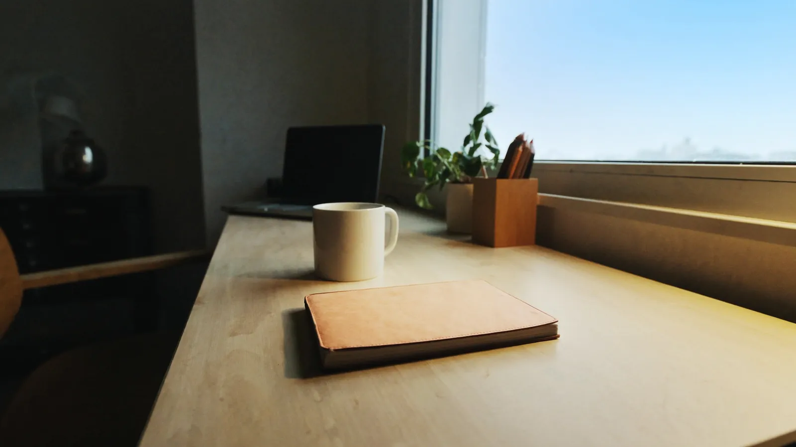 A closed laptop and small potted plant on a sunlit home office desk near a large window