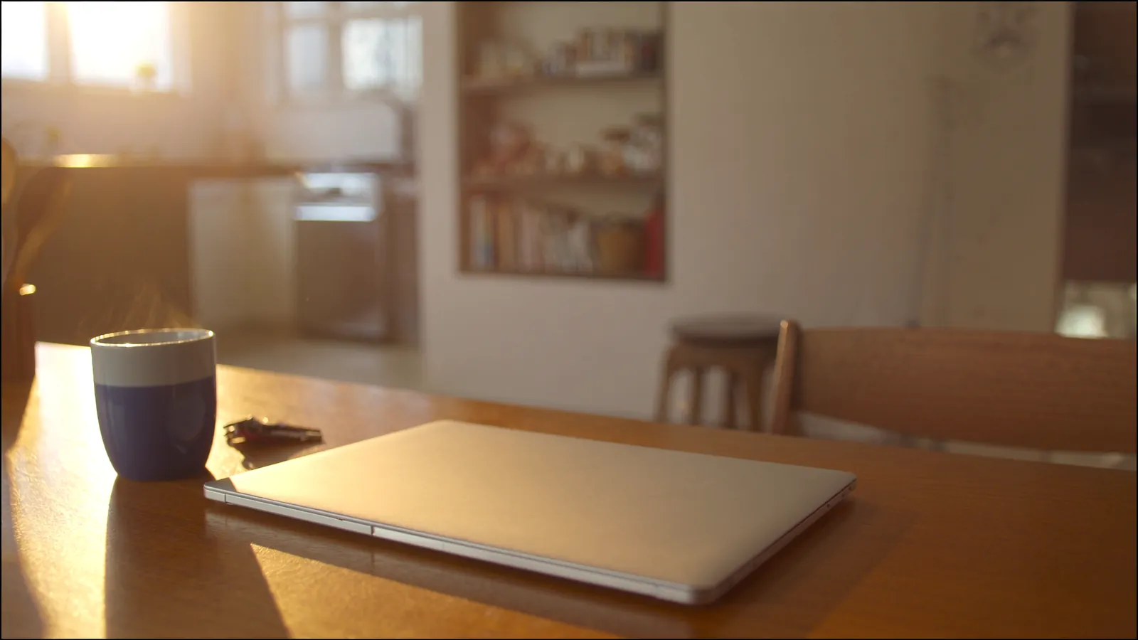 A closed laptop, steaming coffee mug, and house keys on a sunlit wooden kitchen table in the warm morning light of a home interior